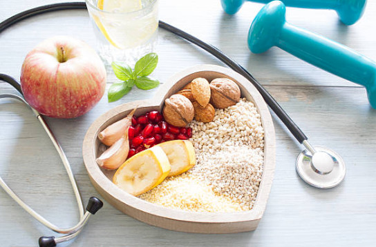 A stethoscope, a wooden heart filled with heart-healthy foods like bananas, nuts, and fruits, alongside handheld weights, depicting a holistic approach to heart health.
